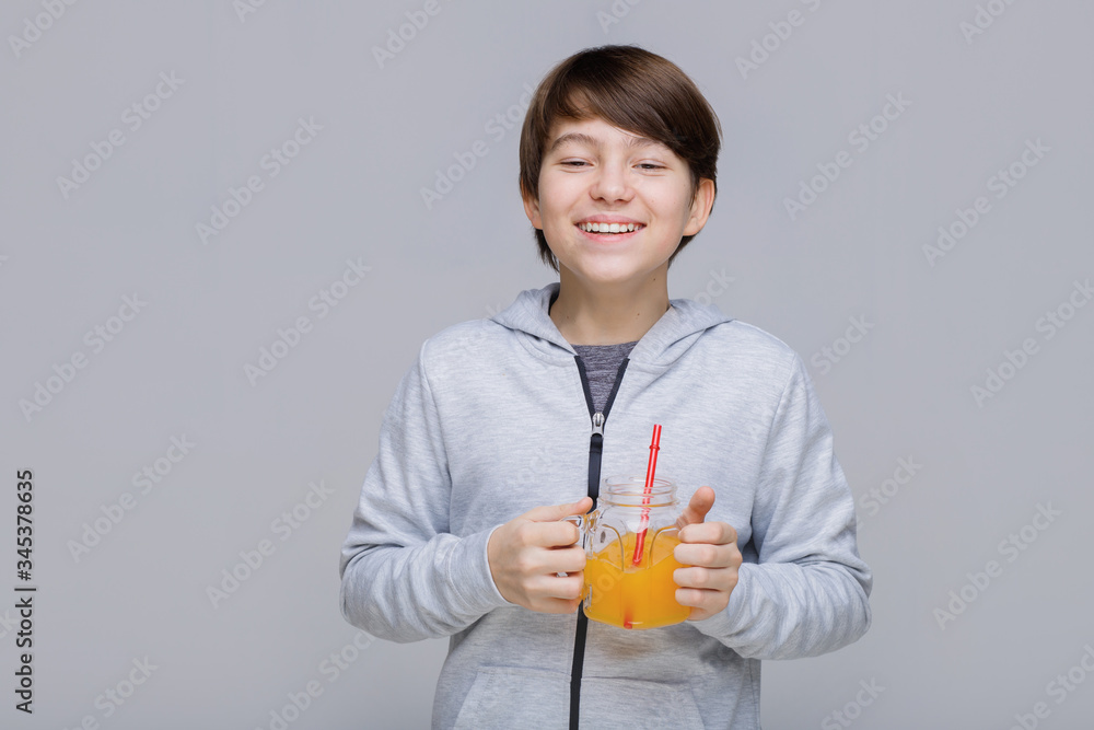 Happy smiling boy drinking fresh juice from glass with a straw. Teenager on gray background. Happy smiling boy drinking fresh juice from glass with a straw. Teenager on gray background.