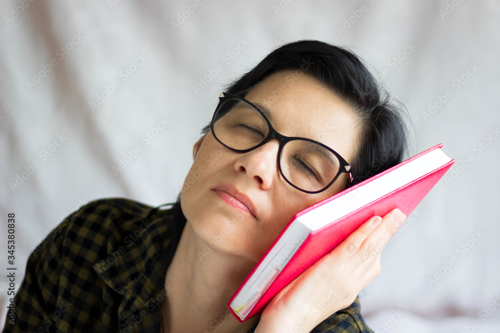 A pretty middle-aged brunette with glasses is dozing with fatigue, her head bowed on a book. The joy of reading and learning in middle age. Emotions. Fatigue A pretty middle-aged brunette with glasses is dozing with fatigue, her head bowed on a book. The joy of reading and learning in middle age. Emotions. Fatigue
