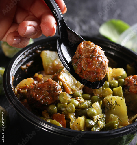 Healthy green food. Stewed cabbage, potatoes, peas and meatballs in a black plastic bowl with a spoon and hand. Green brussels sprouts, spinach on a black background. Background image, copy space