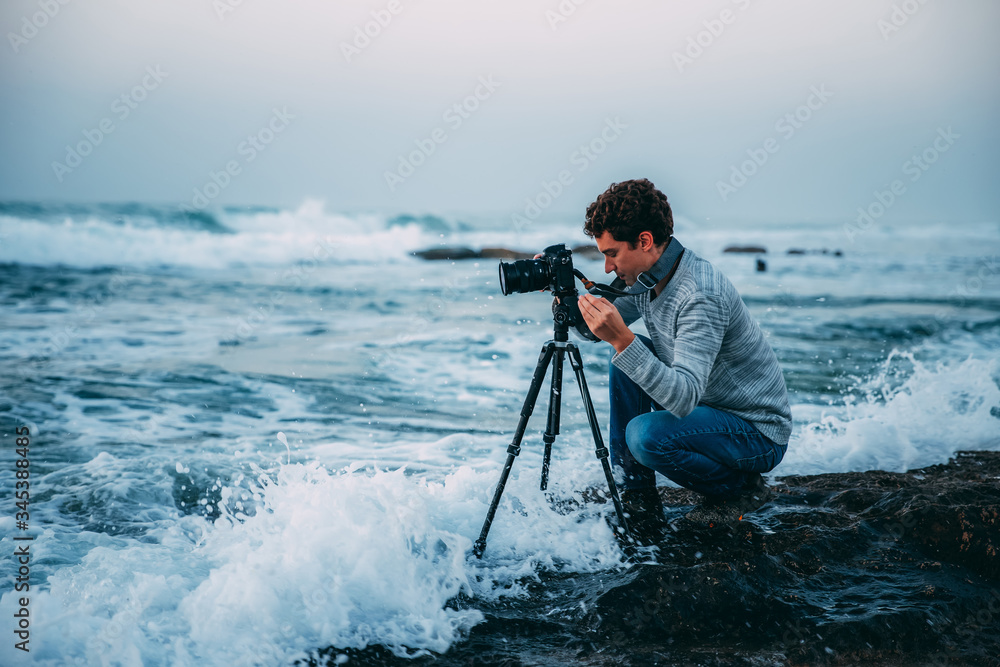 Obraz premium Young handsome photographer with curly hair, a gray sweater and jeans with a tripod and a camera on the beach, drenched in waves. Photography in Israel. Copy space. Shtorm on Mediterranean Sea