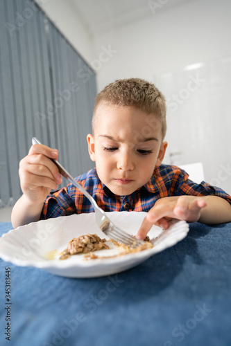 Portrait of small caucasian boy little child kid sitting by the table eating having lunch or dinner holding fork making faces gesture front view at home