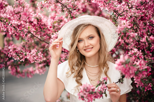 Happy smiling elegant lady wearing white wicker hat, pearl jewelries, dress, posing in pink spring blossom trees. Copy, empty space for text