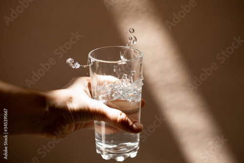 female hand in a ray of sunlight holds a glass with cool fresh clear water with splashes and waves. Art object, the concept of a healthy lifestyle, naturalness, proper nutrition and beauty.