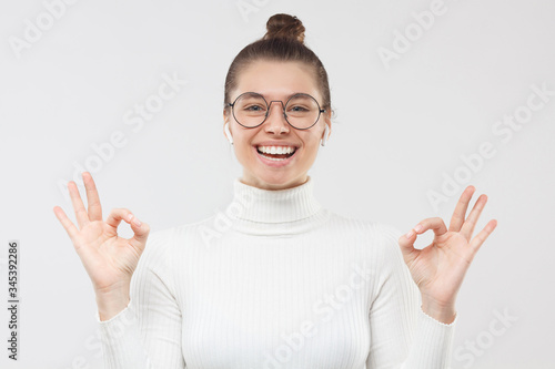 Foto OK! Young happy girl in round glasses and white sweater, showing okay gesture wi