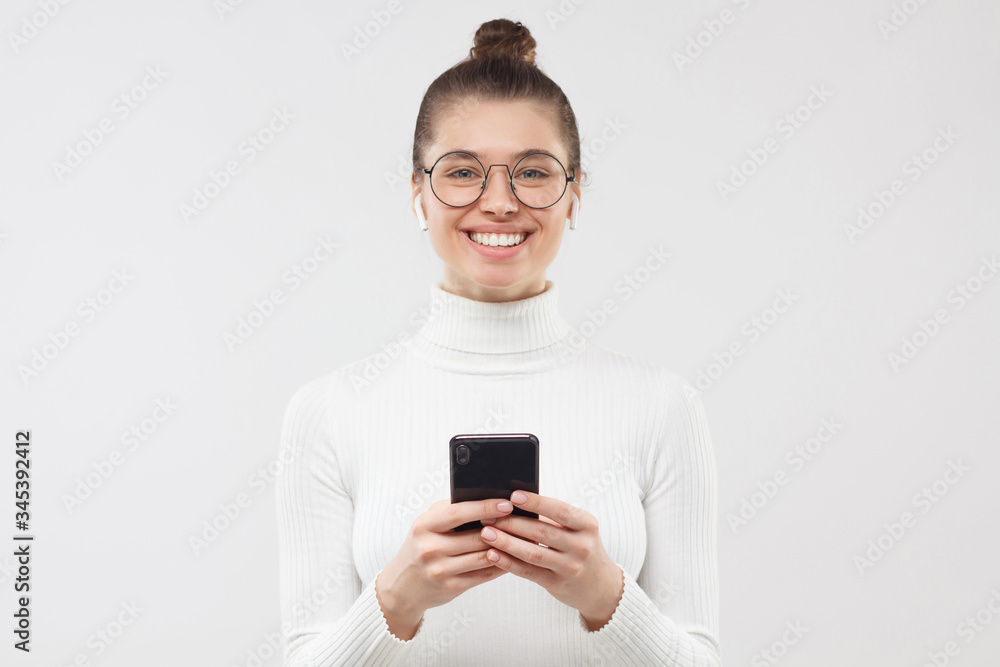 Young girl looking at camera with happy smile, holding phone in hands for communicating with friends, feeling relaxed and positive, isolated on gray background Young girl looking at camera with happy smile, holding phone in hands for communicating with friends, feeling relaxed and positive, isolated on gray background