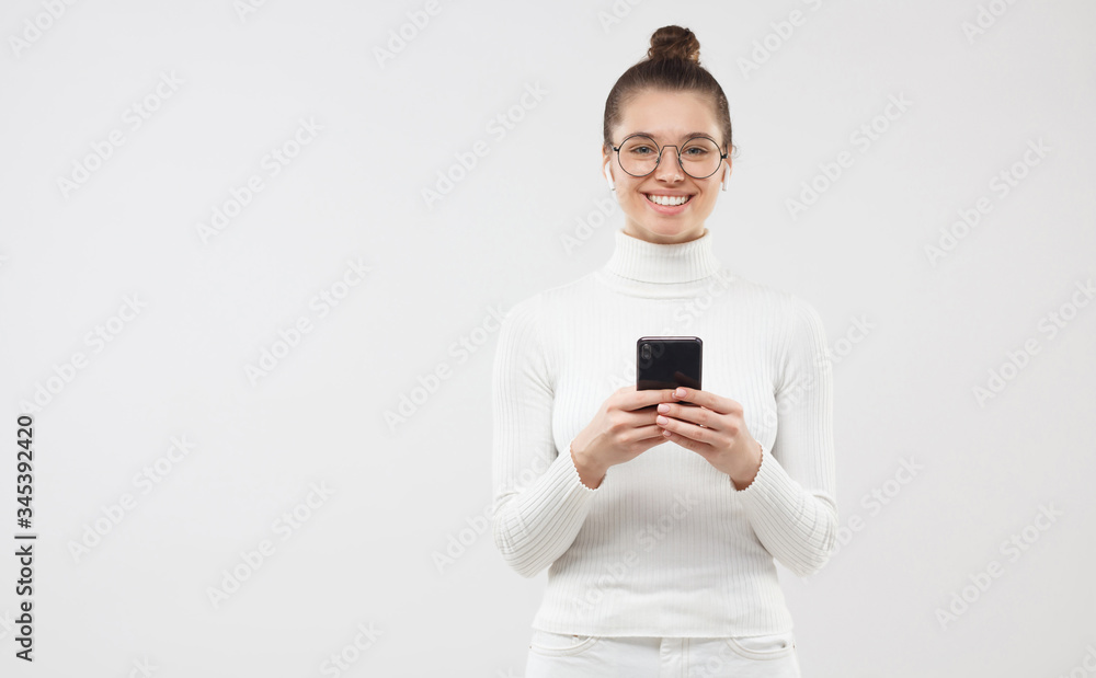 Horizontal banner of young woman holding smart phone in hands, smiling happily at camera, feeling relaxed and calm, isolated on gray background, copy space on left Horizontal banner of young woman holding smart phone in hands, smiling happily at camera, feeling relaxed and calm, isolated on gray background, copy space on left