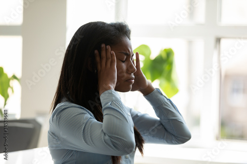 Unhappy african american businesswoman suffering from headache at work at laptop. Diverse female employee stressful touching temples holding head thinking about business problem.