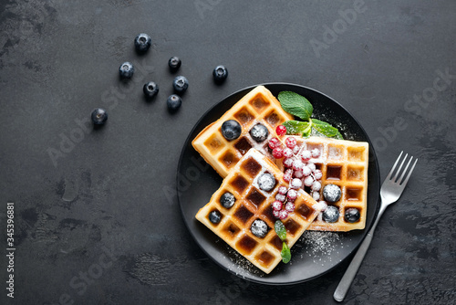 Square belgian waffles with berries and icing sugar on black plate, black background with copy space. Table top view sugar dessert on plate