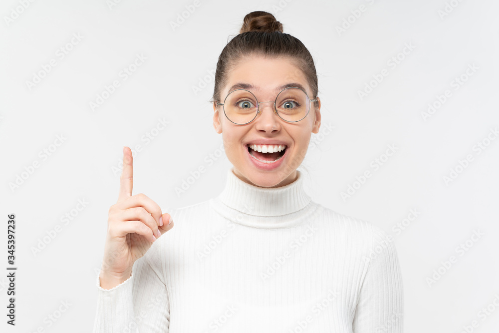 Excited female in eyeglasses pointing up with finger as if she got new idea, feeling happy about good result, smiling, isolated on gray background Excited female in eyeglasses pointing up with finger as if she got new idea, feeling happy about good result, smiling, isolated on gray background