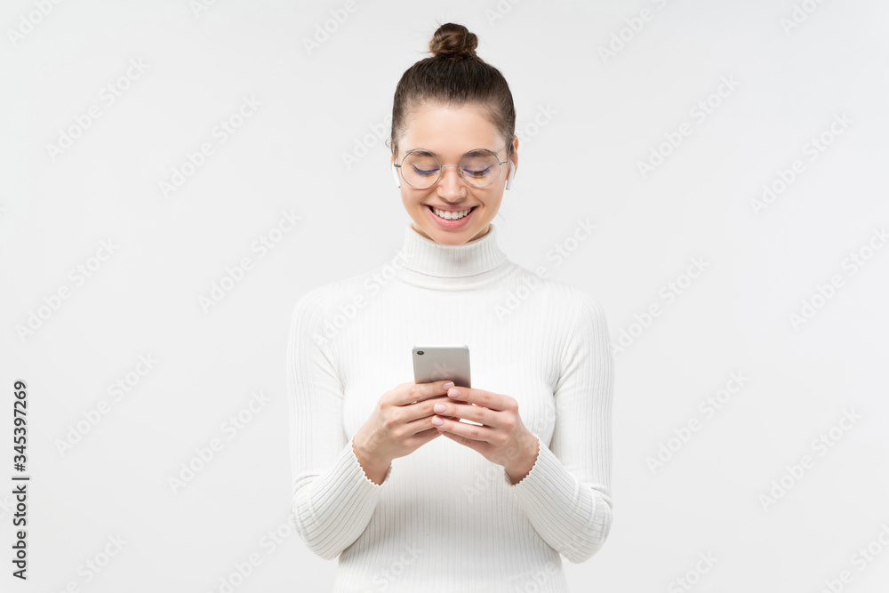 Young woman in glasses, holding smartphone in hands, exchanging messages with friends with happy smile, isolated on gray background Young woman in glasses, holding smartphone in hands, exchanging messages with friends with happy smile, isolated on gray background
