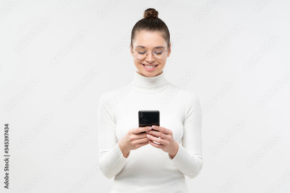Young smiling female student in round eyeglasses standing in front of camera, holding smartphone in both hands, reading text messages, isolated on gray background