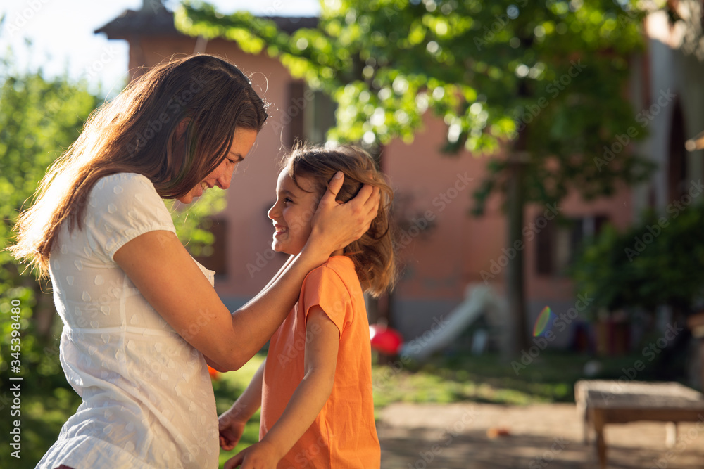 Naklejka premium Authentic shot of happy little daughter and young mother are enjoying their time together outside their house on a sunny day. Concept: family, motherhood,life, happiness, childhood, authenticity