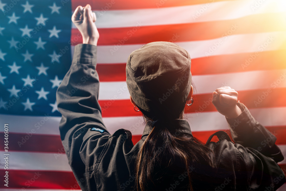 Independence Day. A female soldier rejoices, raising her hands to the top, against the background of the American flag. Rear view. The concept of the American national holidays and patriotism
