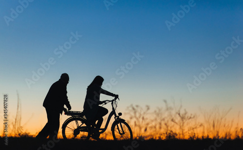 Silhouette of a girl and man on the e-bike or electric bicycle on the sunset background. Country style, transportation in the village. Copy space.Travel, father and daughter.