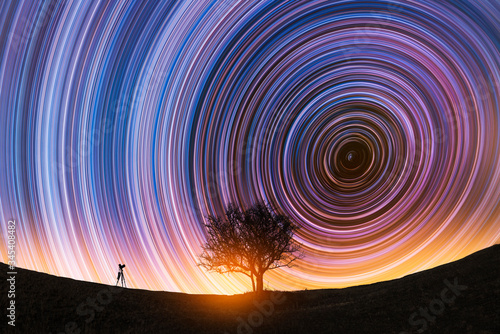 Fotografi Beautiful night landscape, The camera on the tripod and tree silhouette on the hill under the  colorful star trails on the sky