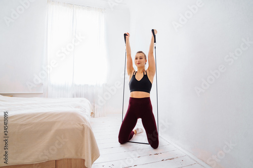 Muscular, attractive, beautiful blonde fitness trainer doing home exercises with a black expander. She crouching and kicking up, smiling. Woman exerting a lot of strength to make her body perfect.