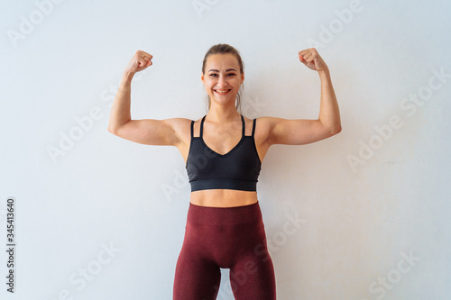 Attractive, muscular fitness girl sits against a white wall with a happy, smiling face and shows her freedom, a well-trained muscular figure and muscles in her arms