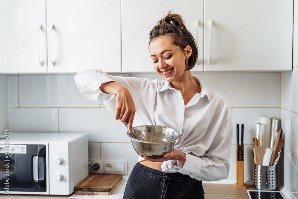 Beautiful, attractive, smiling young woman kneading dough with whisk for banana pancakes in steel dishes in a kitchen with white, light interior. Monochrome photo without bright accents Beautiful, attractive, smiling young woman kneading dough with whisk for banana pancakes in steel dishes in a kitchen with white, light interior. Monochrome photo without bright accents