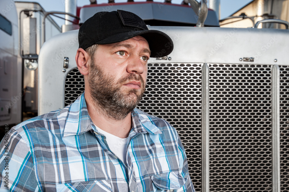 Worried semi truck driver wearing plaid shirt and black baseball hat sits  in front of big