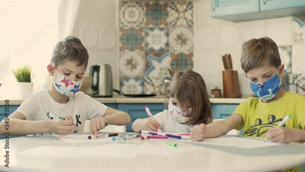 Two Boys And Their Little Sister In Multicolored Medical Masks Draw At The Table In The Kitchen. Two Boys And Their Little Sister In Multicolored Medical Masks Draw At The Table In The Kitchen.