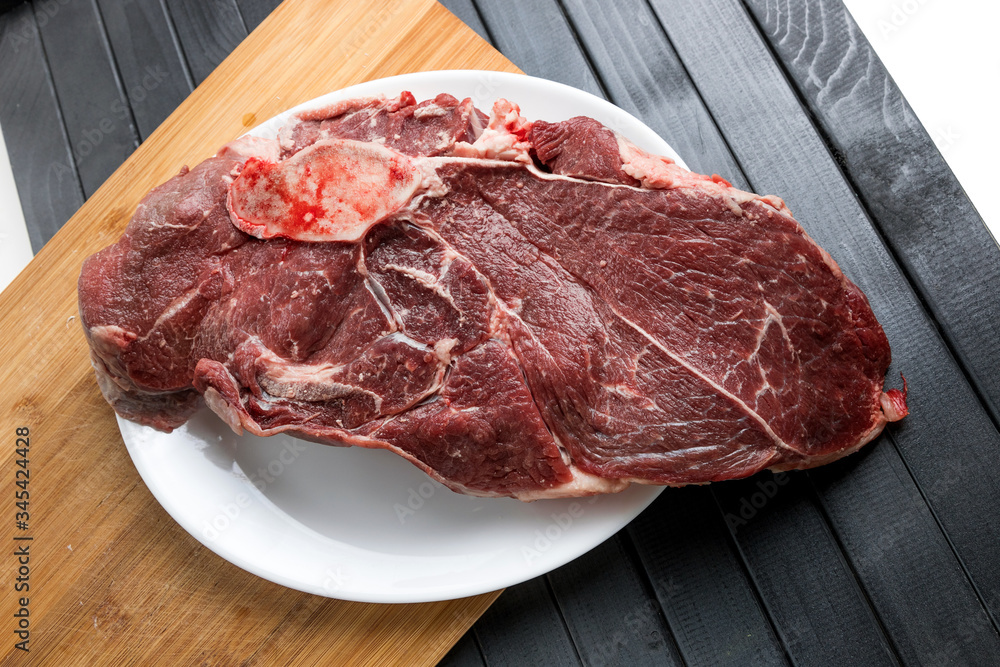 Fototapeta premium A piece of fresh meat on a white plate. Beef steak. Meat cutting. A wooden cutting Board, on a dark table.