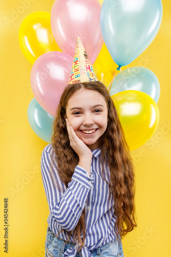 Cute girl with colorful inflatable ballons isolated on a yellow background, little beautiful girl wearing birthday cap, smiling with happy face