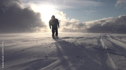 Silhouette of man in black snowmobile overall and helmet, he walking towards Sun shining though heavy clouds in mountains, poor snow visibility, high speed snow drift conditions