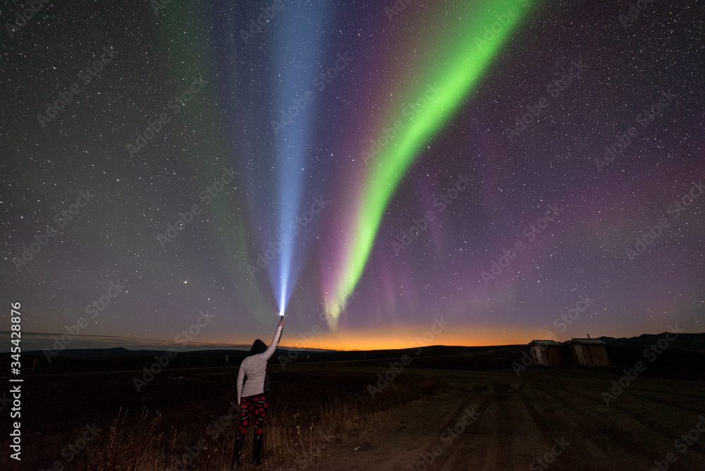 Woman holding flashlight, torch up beside northern lights, aurora borealis band in sky with sunset stars landscape at Arctic Circle, Yukon Territory, Canada. 