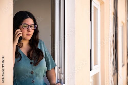Brunette spanish girl dressed on a business serius look talking on the phone at the window from her apartment during the afternoon in Palma de Mallorca, Spain during Coronavirus confinement