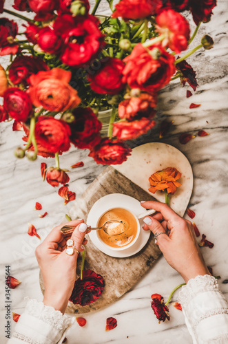 Flat-lay of womans hand holding cup of black espresso coffee and bouquet of red ranunculus flowers over serving board and white marble table background, top view. Seasonal coffee break concept