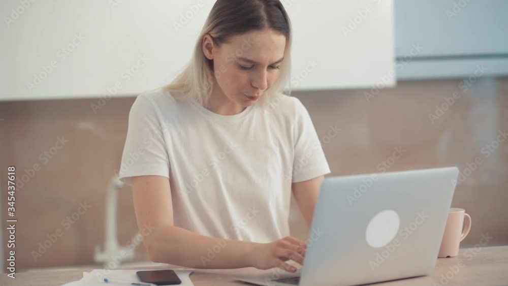 The young girl is concentrated on working in a laptop and distracted by a smartphone. Bright spacious room. The young girl is concentrated on working in a laptop and distracted by a smartphone. Bright spacious room.