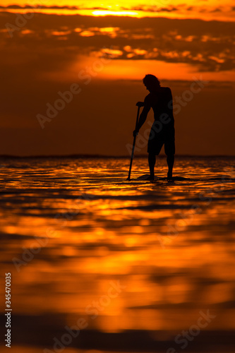 surfer rides by paddle board (S.U.P.) in the ocean against the background of a large disk of the setting sun © ohrim