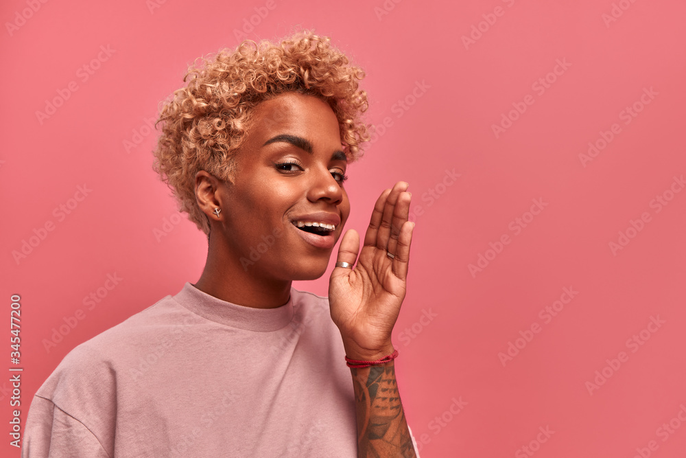 Attention. Close up portrait of cheerful young African American woman with blond hair is holding hand near her open mouth and yell about great sale or discounts, on pink background. Facial expression. Attention. Close up portrait of cheerful young African American woman with blond hair is holding hand near her open mouth and yell about great sale or discounts, on pink background. Facial expression.
