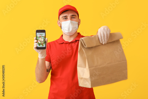Male courier of food delivery company in medical mask and with mobile phone on color background