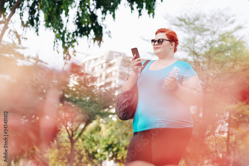 Smiling plus size woman reading text mesage from friend of smartphone when walking outdoors photo