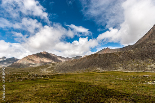 Mountain landscape. Mountain ranges with a green slope in front of them. Blue sky with white clouds. Shadows from the clouds on the mountains. Horizontal.