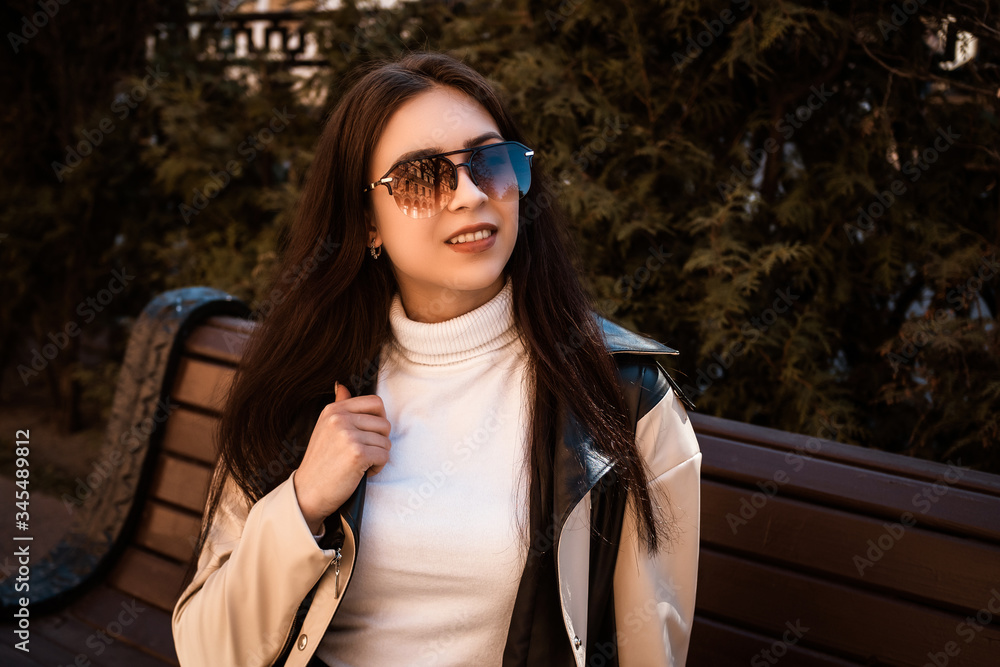 Young woman wearing sweater, sitting on the bench in the city street . Young female student learning outside. In a black and white coat, white sweater, black blouse. In sunglasses