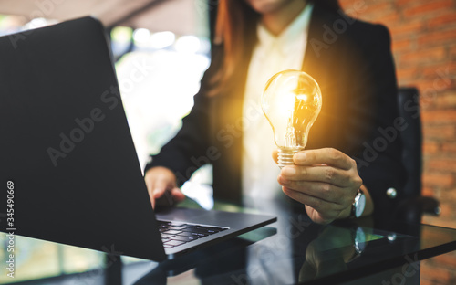 Closeup image of a businesswoman holding a glowing light bulb while working on laptop computer in office