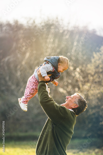 Canvas Print Grandfather proudly rising his granddaugther above his head while enjoying sunny day outdoors