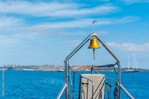 A view of the bell on the stern of the ship in the background blurred the blue sea the coast and some boats that leave the island towards the open sea on a sunny summer day, in Sardinia Italy 
