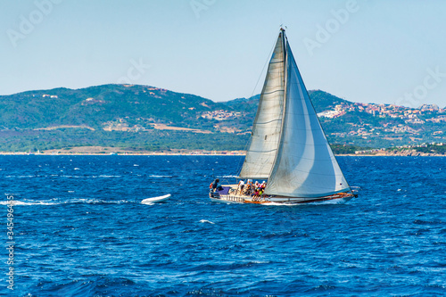 A view of a sailboat while sailing tilted by the wind in the Mediterranean sea with the coast in the background on a day with the sun on summer, in Sardinia Italy 
