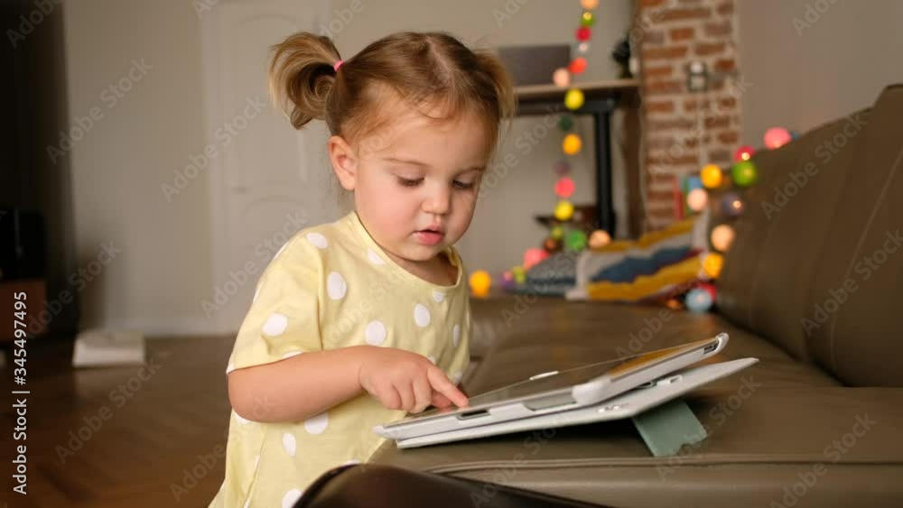 Cute little girl playing game on tablet while resting on blurred background of cozy living room at home Cute little girl playing game on tablet while resting on blurred background of cozy living room at home