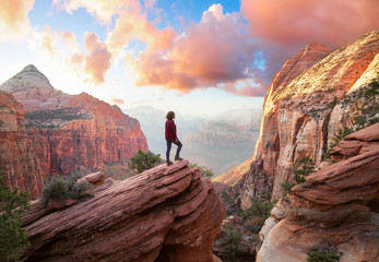 Plexiglas schilderij Beige Adventurous Woman at the edge of a cliff is looking at a beautiful landscape view in the Canyon during a vibrant sunset. Taken in Zion National Park, Utah, United States. Sky Composite.  © edb3_16