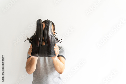 Young caucasian man hidding his face shows his new elegant brown shoes on white wall background,copy space.