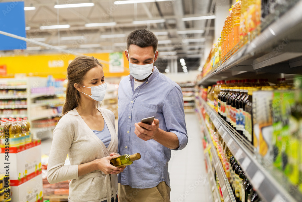 food shopping, consumerism and pandemic concept - happy couple wearing face protective medical masks for protection from virus disease with smartphone buying olive oil at grocery store or supermarket