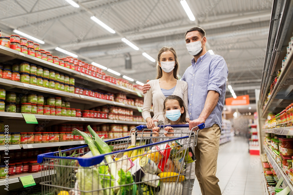 sale, family and pandemic concept - happy mother, father and little daughter wearing face protective medical masks for protection from virus disease with shopping cart buying food at supermarket
