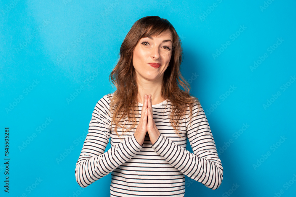 Portrait of young friendly woman with essence in a casual t-shirt makes prayer gesture on an isolated blue background. Emotional face. To ask for something from the universe or from God, make a wish Portrait of young friendly woman with essence in a casual t-shirt makes prayer gesture on an isolated blue background. Emotional face. To ask for something from the universe or from God, make a wish