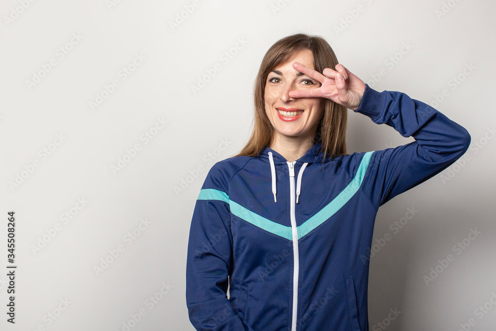 smiling young woman in a 90s sports jacket and showing a V-sign near her eye on a light background. Concept of gestures, style, fashion, sport smiling young woman in a 90s sports jacket and showing a V-sign near her eye on a light background. Concept of gestures, style, fashion, sport