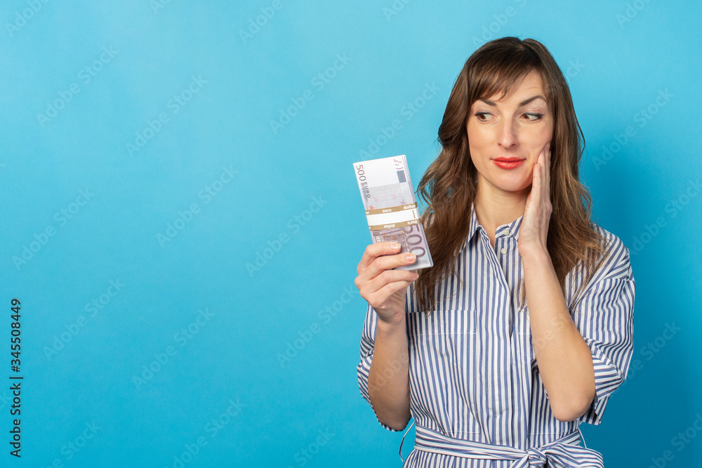 Portrait of a young friendly woman with a smile in a dress holding a bundle of money in her hands and looking at her on an isolated blue background. Emotional face. Concept of wealth, win, credit