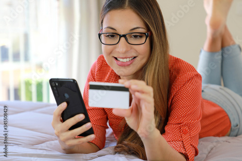 Young confident pretty woman lying on bed reads and enters credit card number for shopping online directly on smartphone from home